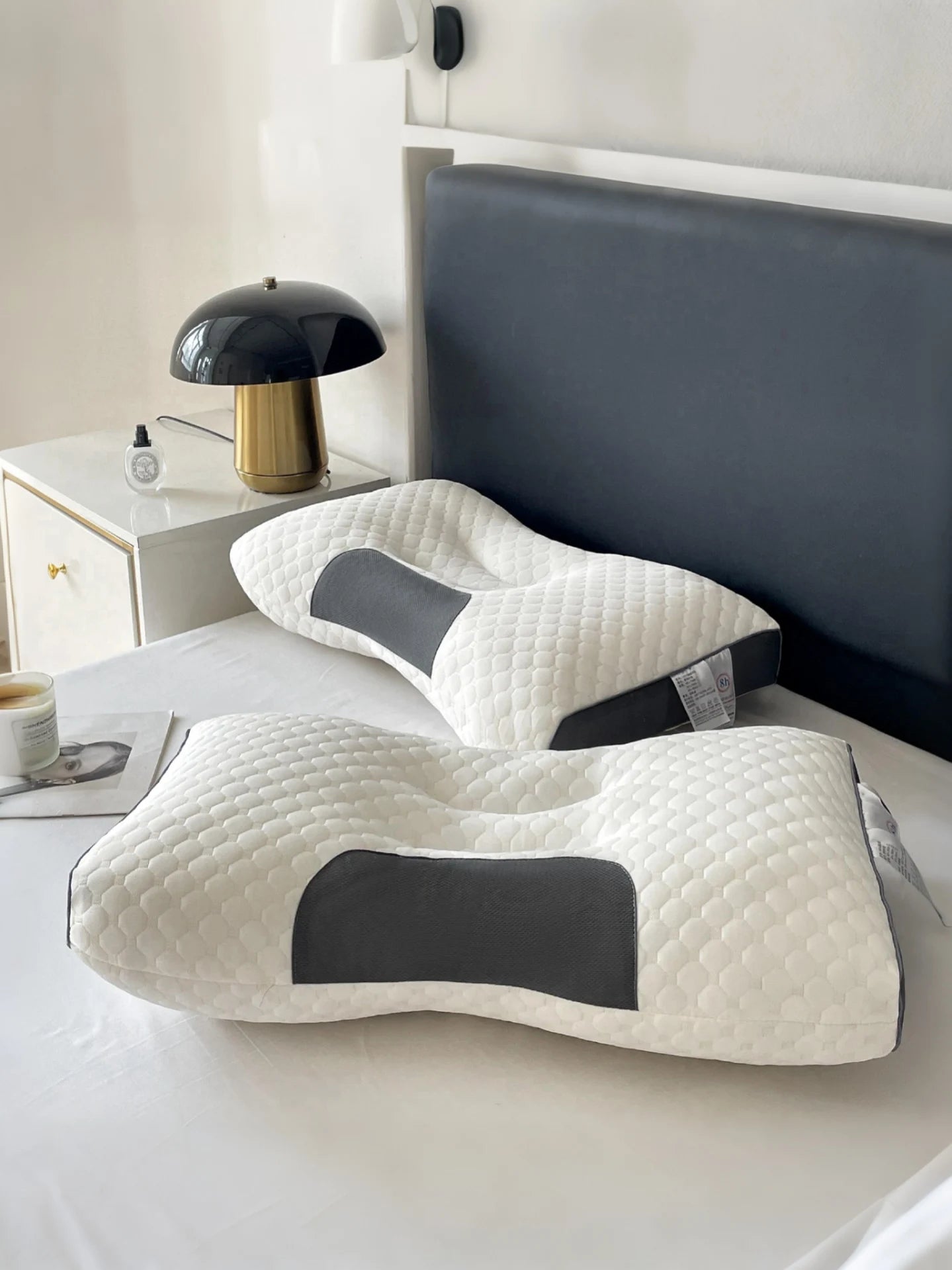 Two ergonomic pillows on a bed with a nightstand and lamp in the background.